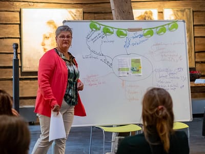 Een vrouw staat bij een whiteboard vol gekleurde notities en moedigt iedereen aan tijd voor jezelf te nemen, in een rustieke kamer.