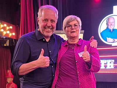 Een man en vrouw staan samen op het podium, lachen, duimen omhoog, vol energie. Op de achtergrond lampen en een groot scherm.