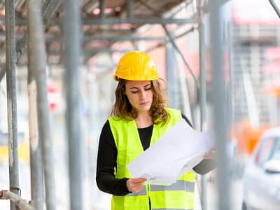 Een vrouw met een gele helm en reflecterend vest bekijkt bouwtekeningen tussen steigers, denkend: wat wil ik verder met mijn leven?