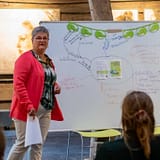 Een vrouw staat bij een whiteboard vol gekleurde notities en moedigt iedereen aan tijd voor jezelf te nemen, in een rustieke kamer.