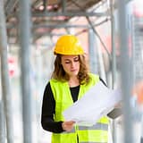 Een vrouw met een gele helm en reflecterend vest bekijkt bouwtekeningen tussen steigers, denkend: wat wil ik verder met mijn leven?