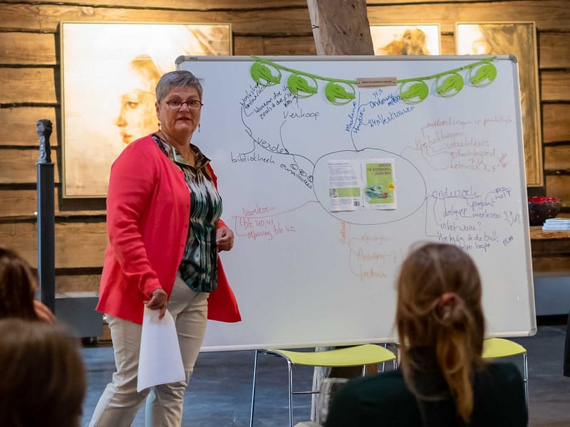 Een vrouw staat bij een whiteboard vol gekleurde notities en moedigt iedereen aan tijd voor jezelf te nemen, in een rustieke kamer.
