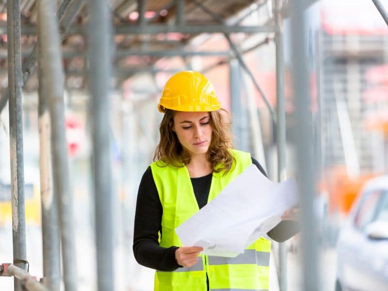 Een vrouw met een gele helm en reflecterend vest bekijkt bouwtekeningen tussen steigers, denkend: wat wil ik verder met mijn leven?