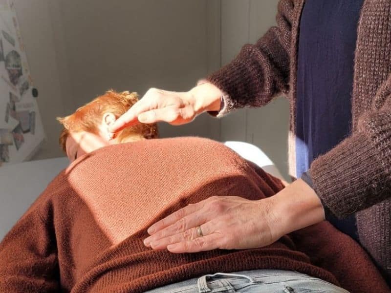 Iemand in een bruine trui krijgt een reiki-behandeling op de rug en nek van een behandelaar.