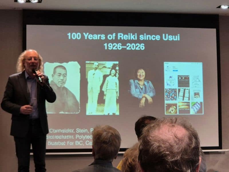 Een man staat achter een lessenaar voor een projectiescherm met de titel 100 Years of Reiki since Usui 1926-2026, waarop historische foto’s en een socialemediaprofiel van Reiki Breda te zien zijn.